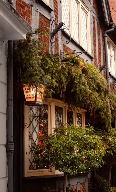 The image depicts the front exterior of a traditional brick house with decorative timber framing. A lush, green hanging plant with dense foliage drapes over a wrought iron bracket attached above a dou