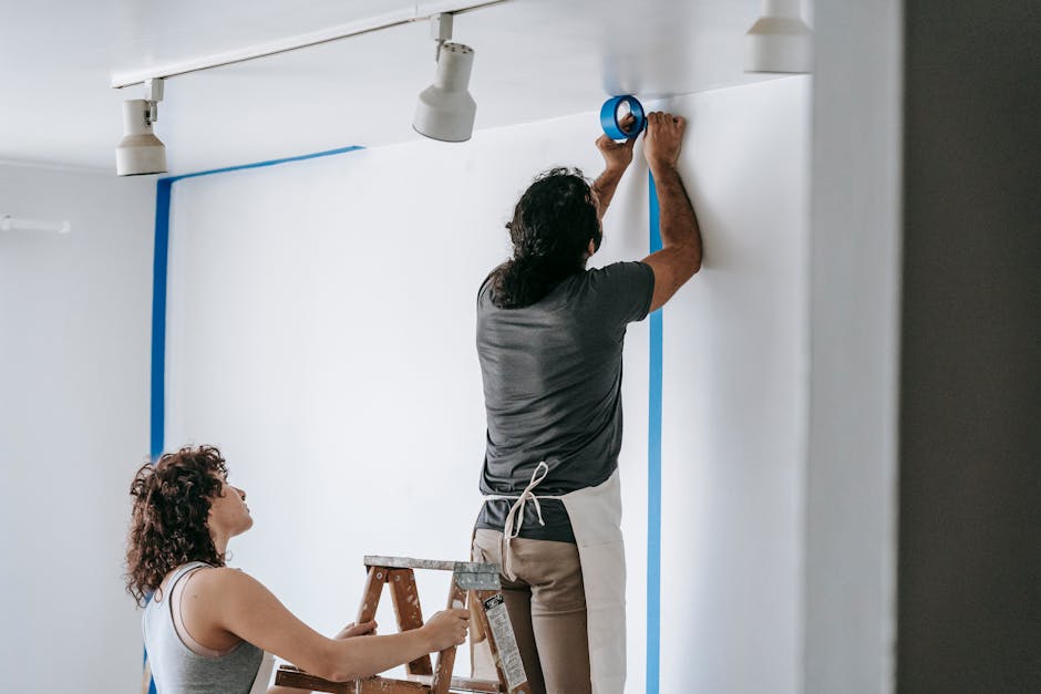A man with long, dark wavy hair wearing a gray T-shirt and beige pants is standing on a small step ladder, applying blue painter's tape along the edges of a white wall to prepare for painting or decor