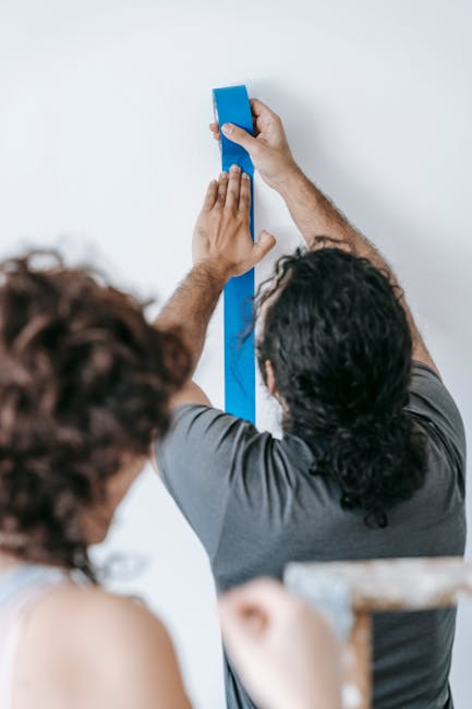 A man with long, dark wavy hair wearing a gray T-shirt and beige pants is standing on a small step ladder, applying blue painter's tape along the edges of a white wall to prepare for painting or decor